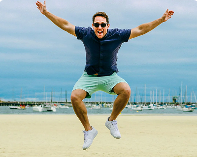 Man wearing light mint green men's shorts made of soft fabric paired with a navy short-sleeve shirt and white sneakers, jumping energetically on a sandy beach with boats and water in the background.
