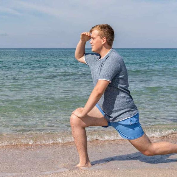 A young man does a lunge on a sandy beach while looking out at the ocean, holding his hand above his eyes to shield from the sun. He wears a gray t-shirt and blue frankster shorts. The sea appears calm under a partly cloudy sky.