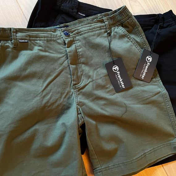 A pair of army green shorts and a pair of black shorts, both with tags attached, are laid out on a wooden surface. The tags display the brand Frankster.