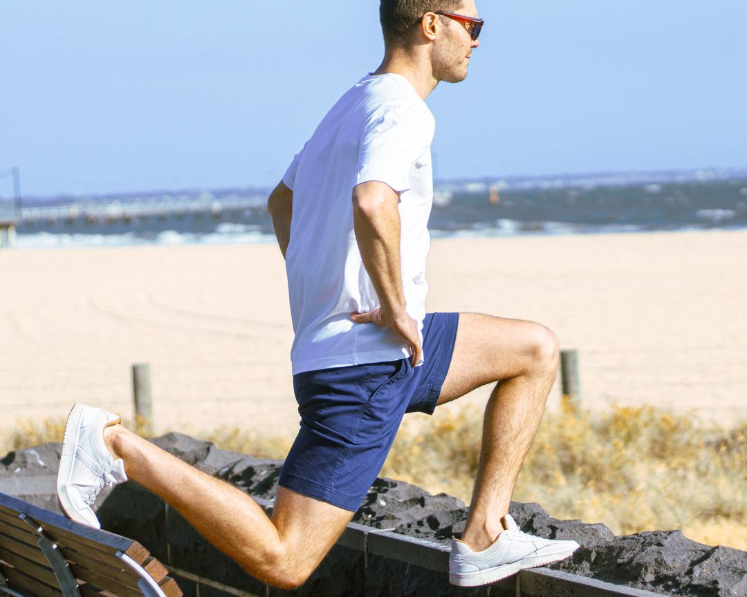 A man wearing sunglasses, a white t-shirt, and light green stretch cotton men's shorts exercises by the beach. He is stretching with one foot resting on a low stone wall and the other leg bent, while holding his hands on his hips. The ocean and sandy beach are visible in the background.