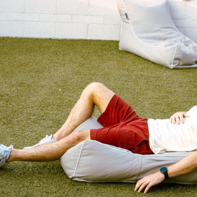A man in a white shirt, red frankster men shorts, and a white cap lies on a wooden bench in the park. He has his right arm resting on his chest and wears reflective sunglasses. The sun is shining brightly, casting shadows, and the grass around is green and lush—a typical Frankster enjoying the day with "The Pioneers.