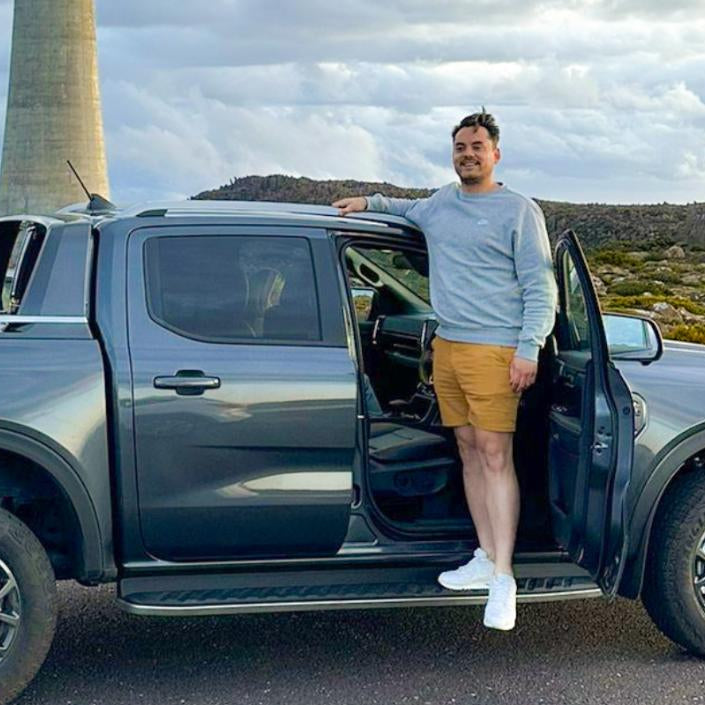 A man in a gray sweatshirt and dark yellow frankster shorts stands on the footboard of a gray pickup truck with the driver's side door open. He is smiling and leaning on the roof of the truck. In the background, there is a large tower and a cloudy sky.