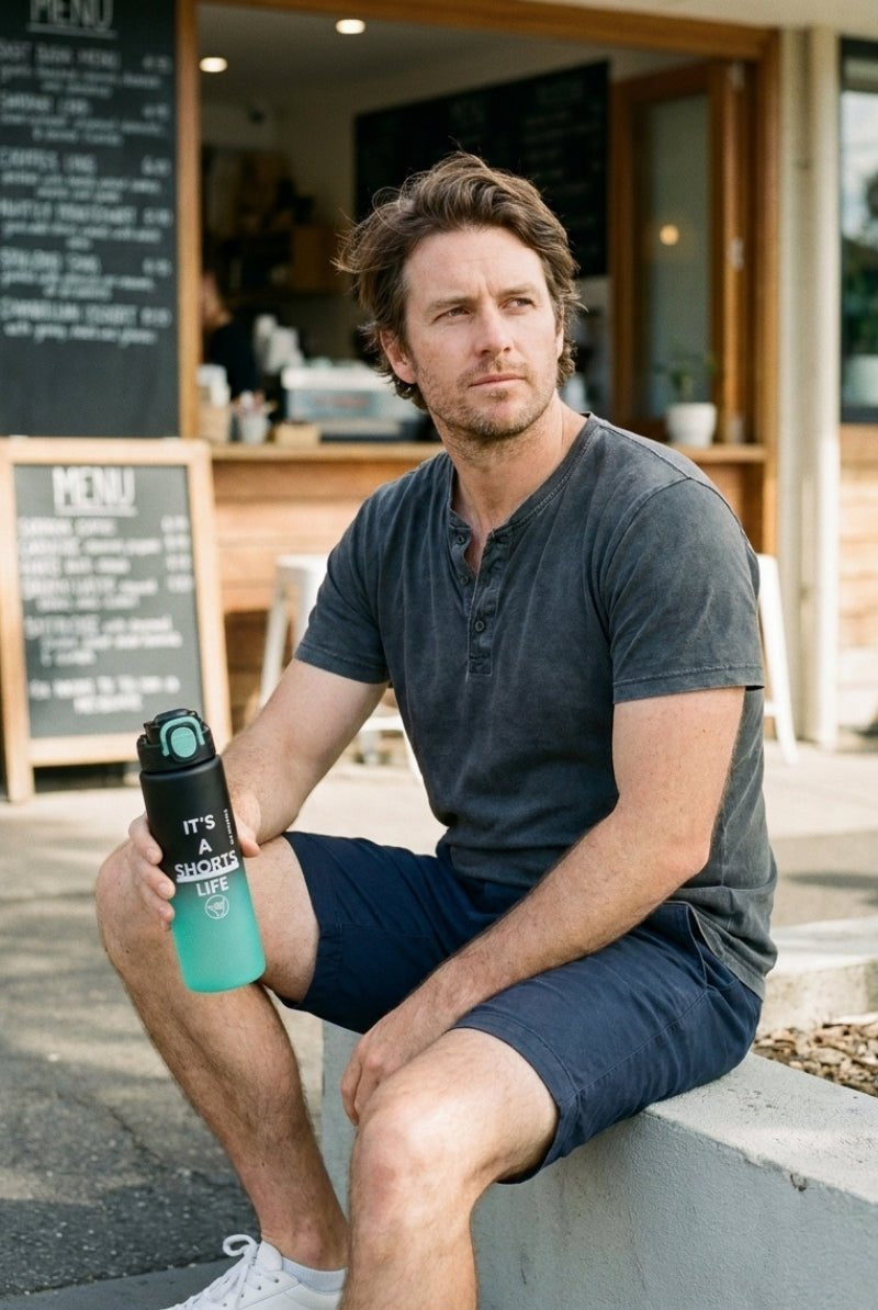 Man wearing Frankster Shorts Life Bottle Green Black men's shorts in a casual dark wash sitting outdoors holding a matching green and black bottle, showcasing comfortable men's shorts for everyday wear.
