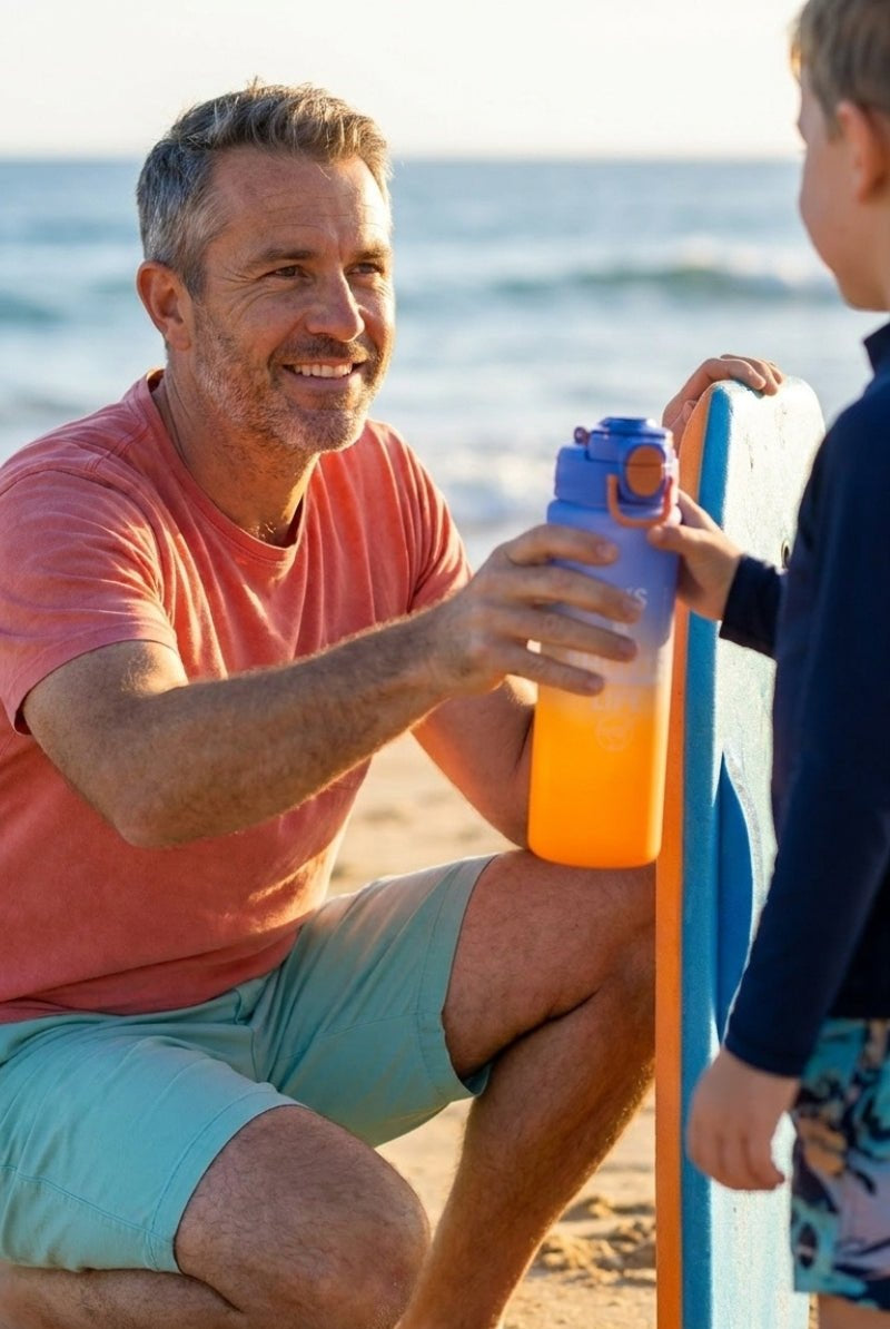 Man wearing light green Frankster men's shorts, handing a vibrant Water Bottle Orange Blue to a child at the beach in a casual, comfortable setting.