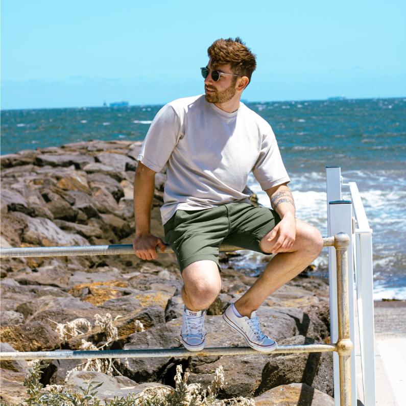 A man wearing a light beige t-shirt, army green frankster shorts, and white sneakers is sitting on a railing near a rocky shoreline. He is looking into the distance with the ocean and waves behind him, under a clear blue sky. He is also wearing sunglasses.