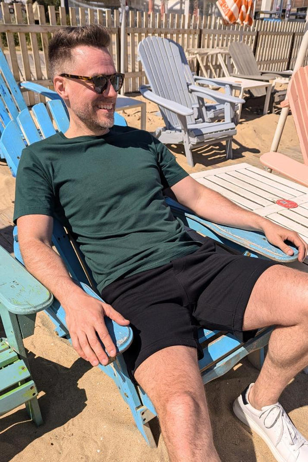 Man wearing sunglasses, dark green t-shirt, black shorts, and white sneakers sitting on a blue wooden chair on sand outdoors