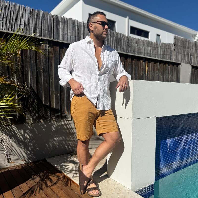 Man wearing mustard yellow men's shorts made from lightweight fabric paired with a loose white linen shirt and dark sunglasses, standing by a pool on a sunny day.