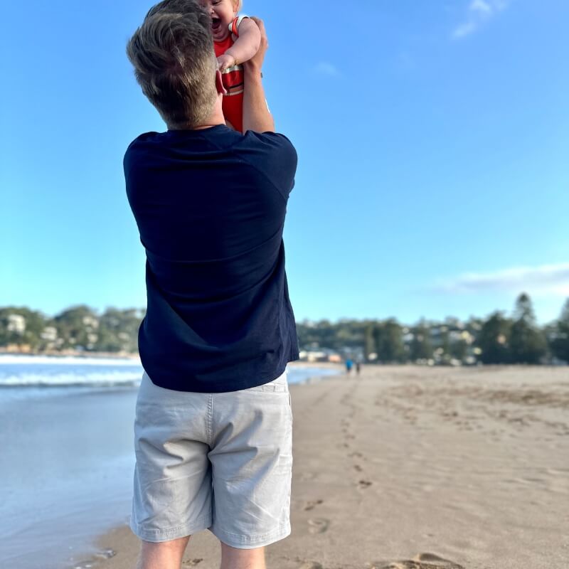 Man wearing light beige men's shorts made from breathable fabric paired with a dark navy shirt, standing on a sandy beach holding a child under bright blue sky.