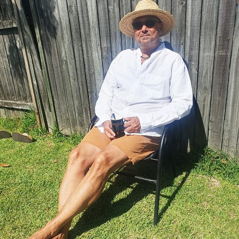 Man sitting outdoors on a black chair wearing warm brown men's shorts made from soft fabric, paired with a loose white long-sleeve shirt and straw hat in bright sunlight.