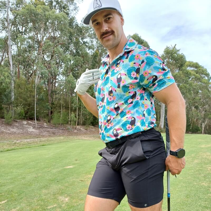 Man wearing black men's shorts made from smooth, stretchy fabric paired with a bright tropical print short-sleeve shirt and a white cap, standing on a golf course.