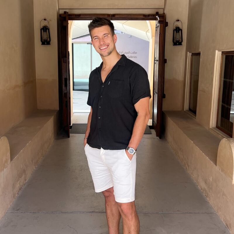 Man standing in an archway wearing casual white cotton men's shorts paired with a short-sleeve black button-up shirt, smiling with hands in pockets.