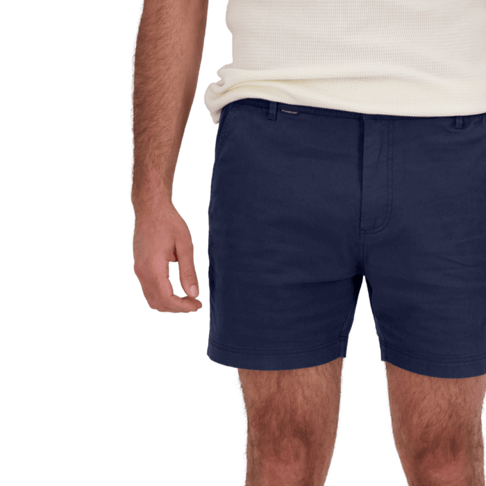 Man wearing navy blue shorts and a cream sleeveless shirt with hand relaxed by side against plain background