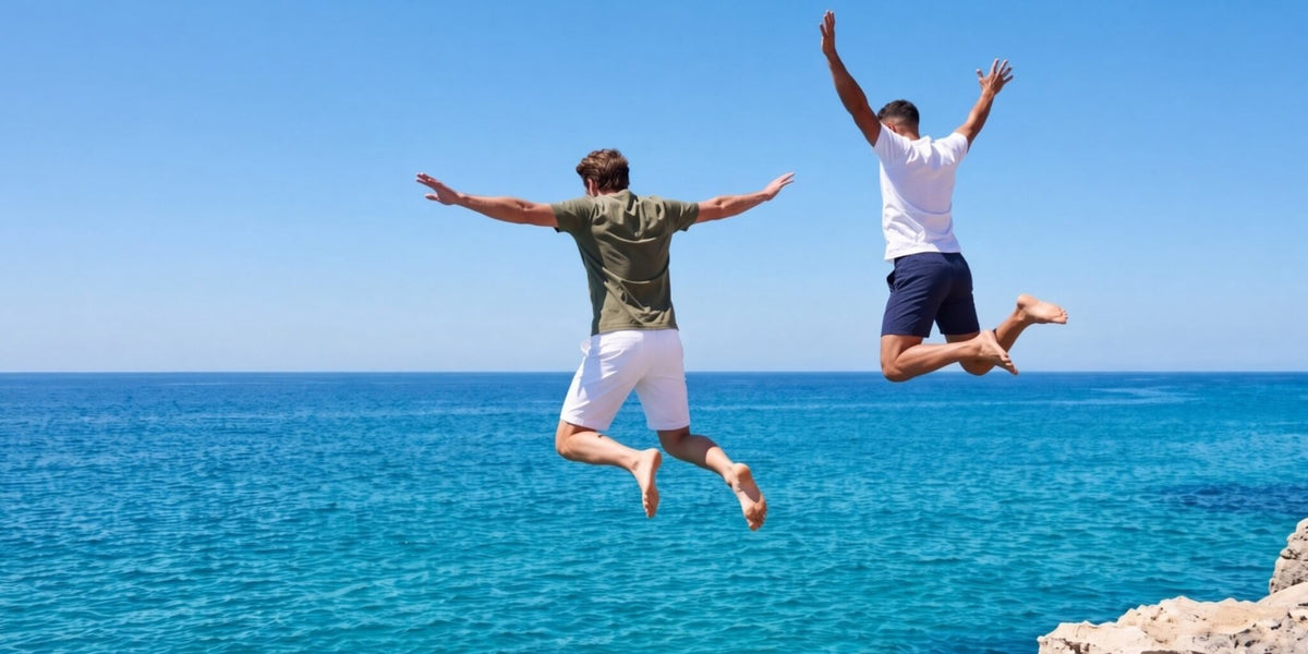 Two men jumping above a bright blue ocean wearing white and navy Men's Shorts with fabric details not visible