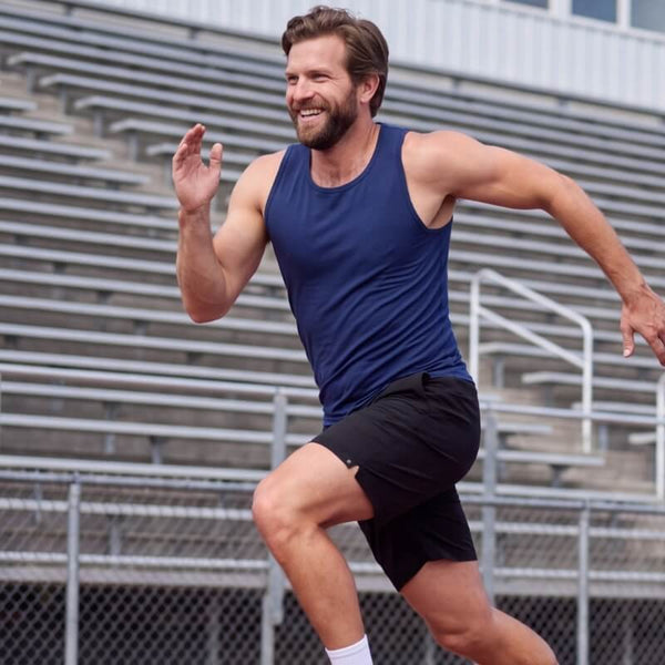 Man running outdoors in black fabric Men's Shorts and a navy tank top beside stadium bleachers