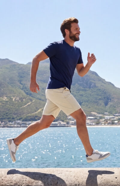 Man running beside a sparkling waterfront in beige Men's Shorts, navy shirt, and white trainers with green mountains behind him