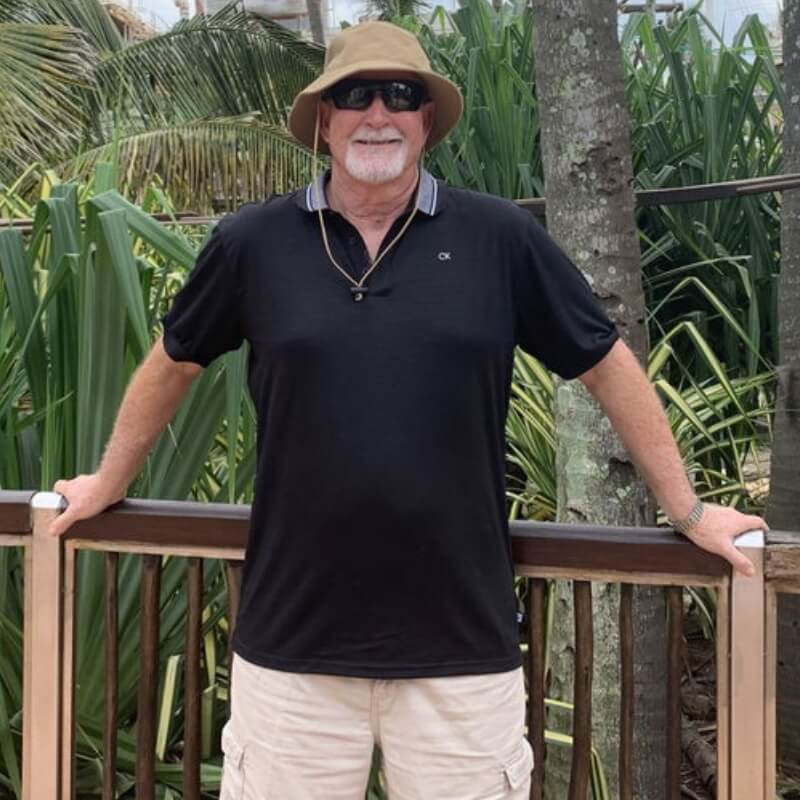 Older man wearing beige Men's Shorts and a black polo shirt outdoors among tropical plants and a wooden railing