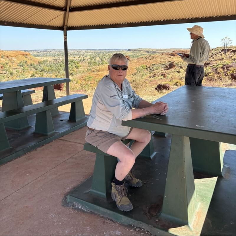 Man wearing khaki Men's Shorts sits at a picnic table beneath a shelter overlooking a rugged Australian landscape