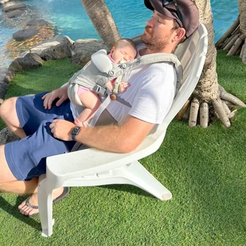 A man wearing blue fabric Men's Shorts sits in a white chair while holding a sleeping baby beside a pool
