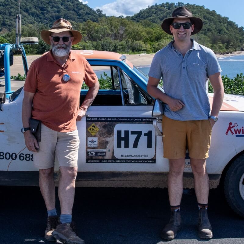 Two men wearing beige and mustard Men's Shorts beside a rally car outdoors, with casual fabric styles visible.
