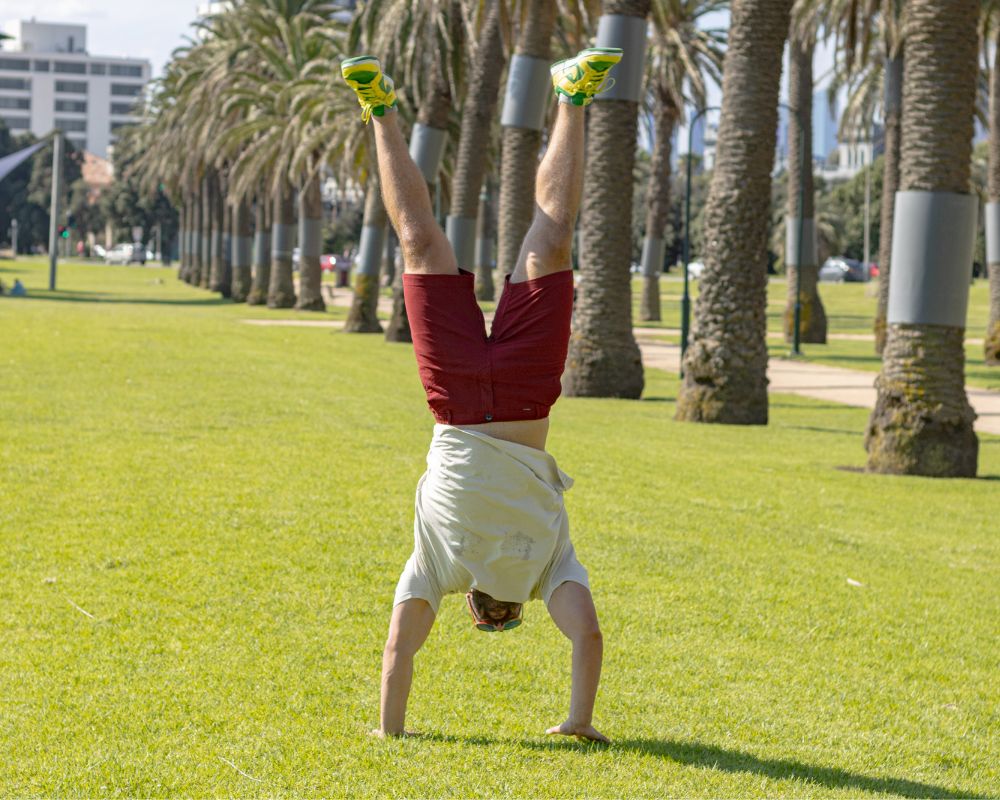 A person in a white shirt and red frankster shorts performs a handstand on a grassy lawn lined with tall palm trees on a sunny day. Buildings are visible in the background.