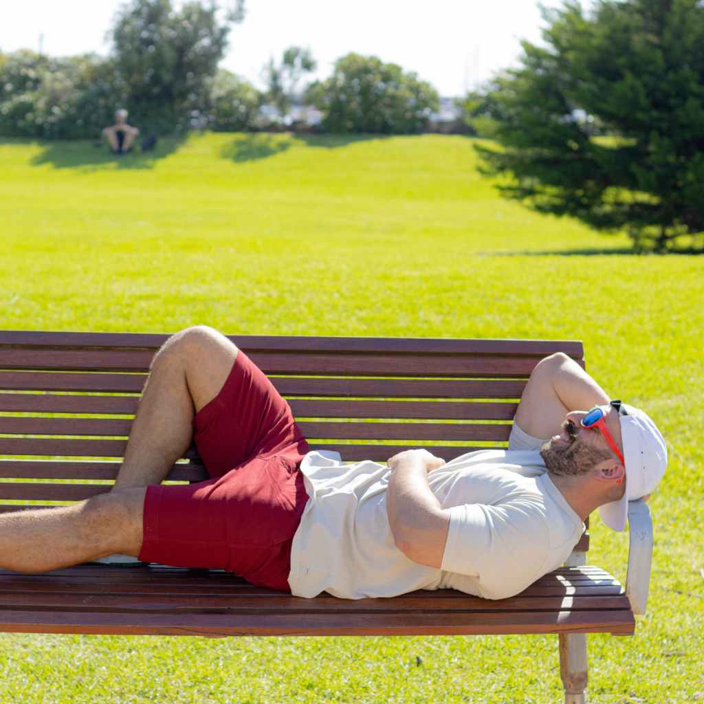 A man in a white shirt, red frankster men shorts, and a white cap lies on a wooden bench in the park. He has his right arm resting on his chest and wears reflective sunglasses. The sun is shining brightly, casting shadows, and the grass around is green and lush—a typical Frankster enjoying the day with "The Pioneers.