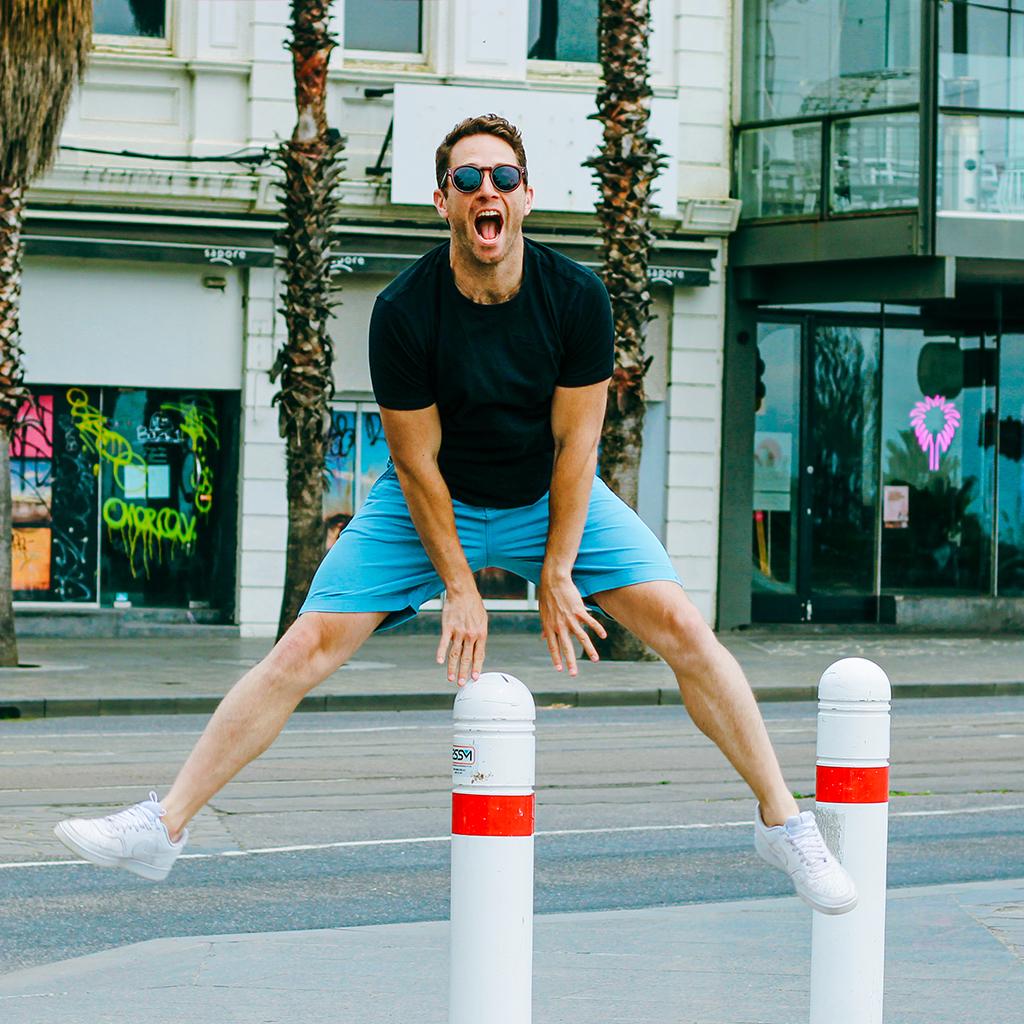 A man in a black t-shirt, blue frankster shorts, and white sneakers is mid-jump, playfully straddling two white posts on a city street. He is wearing sunglasses and smiling energetically. Behind him are palm trees and a building with colorful graffiti on the windows.