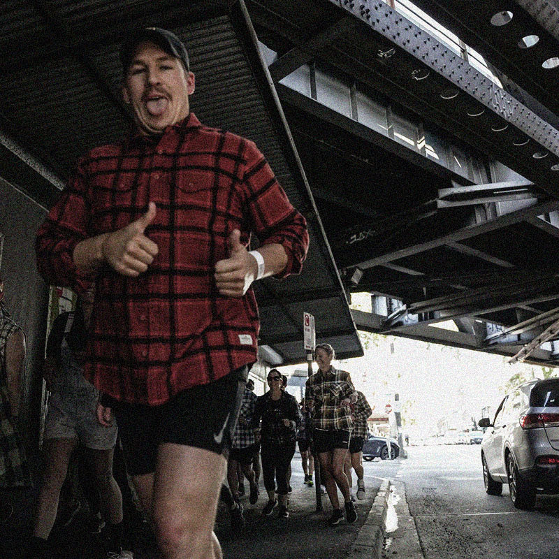 A person wearing a red flannel shirt and black shorts gives two thumbs up while sticking out their tongue. They are walking on a city street under a large metal structure, accompanied by a group of people behind them. Cars are parked on the right side of the street.