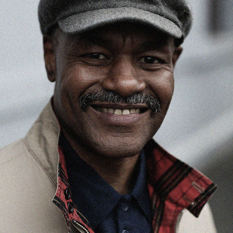 A smiling man with a grey mustache and beard, wearing a grey flat cap and beige jacket with a red-plaid lining. He stands outdoors against an out-of-focus background.