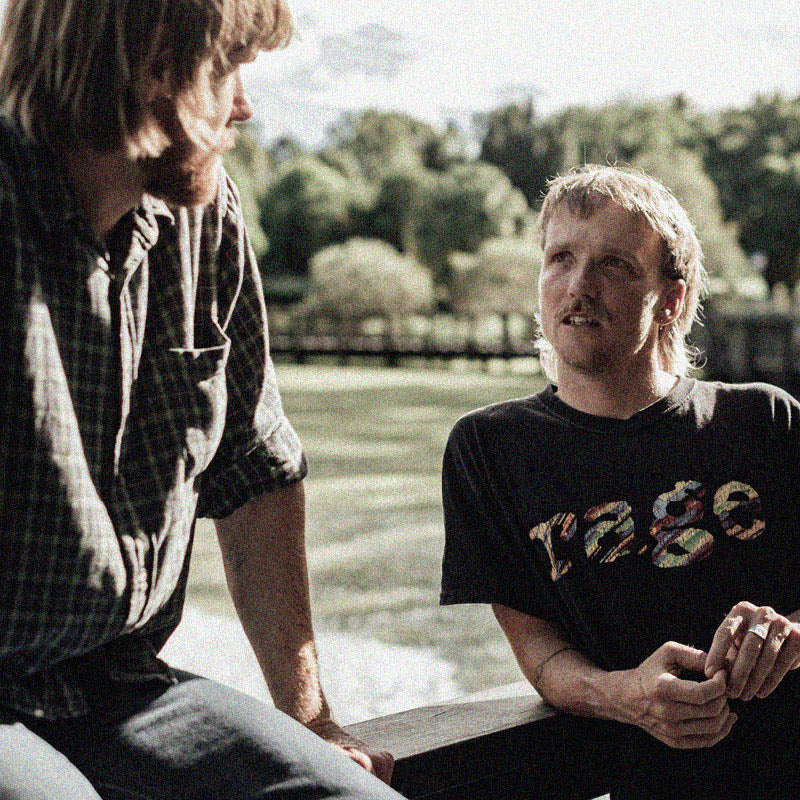 Two men are outdoors, leaning on a wooden railing with a river and trees in the background. The man on the left has shoulder-length hair, a beard, and is wearing a plaid shirt. The man on the right has short hair and a mustache, wearing a black "rage" T-shirt.