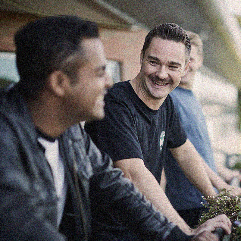 Three men are standing and smiling while leaning against a railing outdoors. The man in the foreground on the left is slightly blurred, while the man in the middle is clearly visible, looking at him and smiling. The third man is in the background, partially visible.