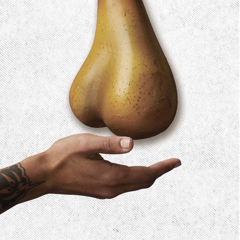 A hand with a tattoo on the wrist is extended upwards, palm facing up, towards a brown object resembling a pear hanging from above. The background is a textured white surface.