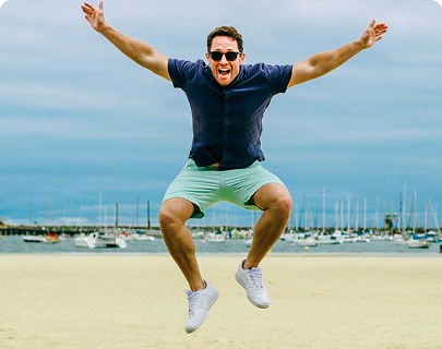 Man wearing light mint green men's shorts made of soft fabric paired with a navy short-sleeve shirt and white sneakers, jumping energetically on a sandy beach with boats and water in the background.