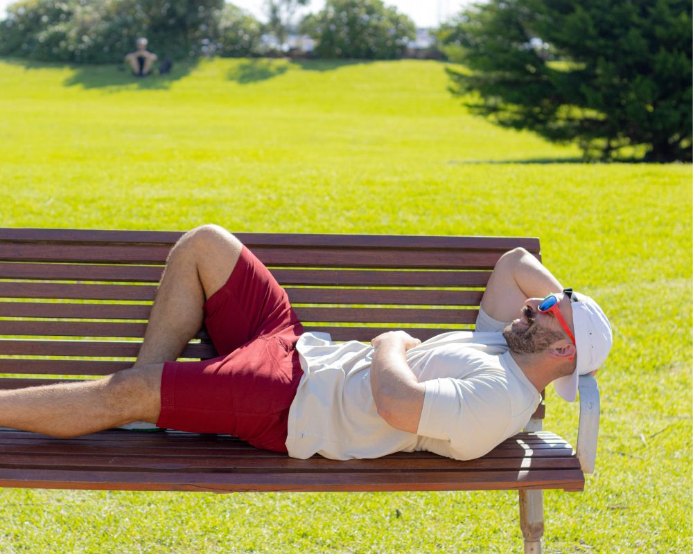 A man in a white shirt, red men shorts, and a white cap lies on a wooden bench in the park. He has his right arm resting on his chest and wears reflective sunglasses. The sun is shining brightly, casting shadows, and the grass around is green and lush—a typical Frankster enjoying the day with "The Pioneers.