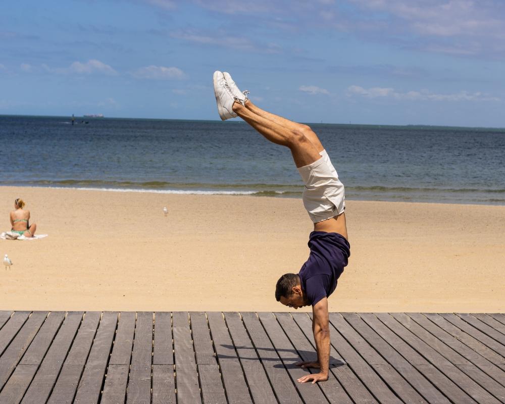 A man in a navy t-shirt and white frankster shorts performs a handstand on a wooden boardwalk with a sandy beach and ocean in the background. A person in swimwear sits on a towel nearby, enjoying the sunny day.