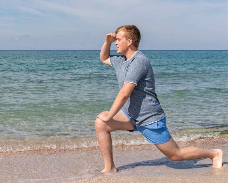 A young man does a lunge on a sandy beach while looking out at the ocean, holding his hand above his eyes to shield from the sun. He wears a gray t-shirt and blue frankster shorts. The sea appears calm under a partly cloudy sky.