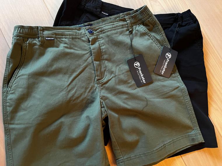 A pair of army green shorts and a pair of black shorts, both with tags attached, are laid out on a wooden surface. The tags display the brand Frankster.