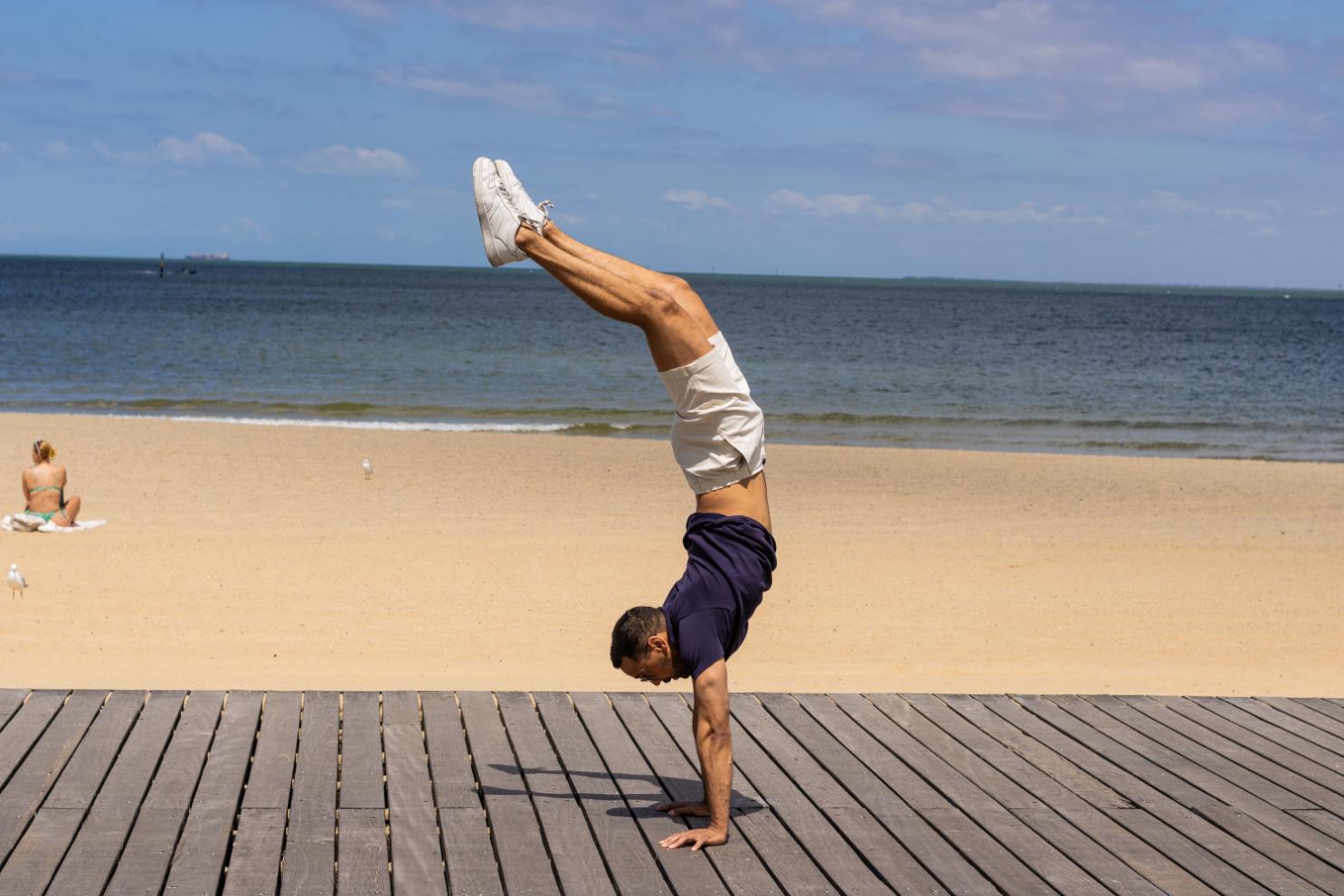 A man in a navy t-shirt and white frankster shorts performs a handstand on a wooden boardwalk with a sandy beach and ocean in the background. A person in swimwear sits on a towel nearby, enjoying the sunny day.