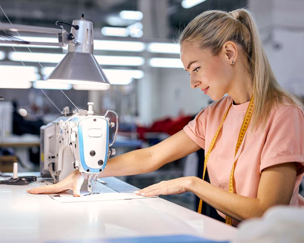 A woman with long blonde hair, wearing a pink shirt and yellow measuring tape around her neck, is focused on using an industrial sewing machine in a brightly lit workshop. She guides fabric smoothly under the needle with both hands.