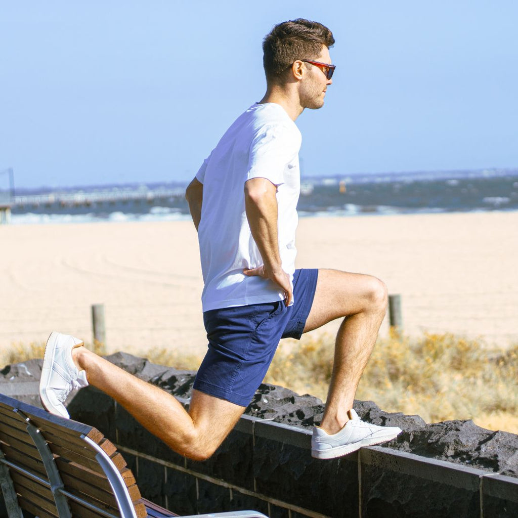 A man wearing sunglasses, a white t-shirt, and light green stretch cotton men's shorts exercises by the beach. He is stretching with one foot resting on a low stone wall and the other leg bent, while holding his hands on his hips. The ocean and sandy beach are visible in the background.