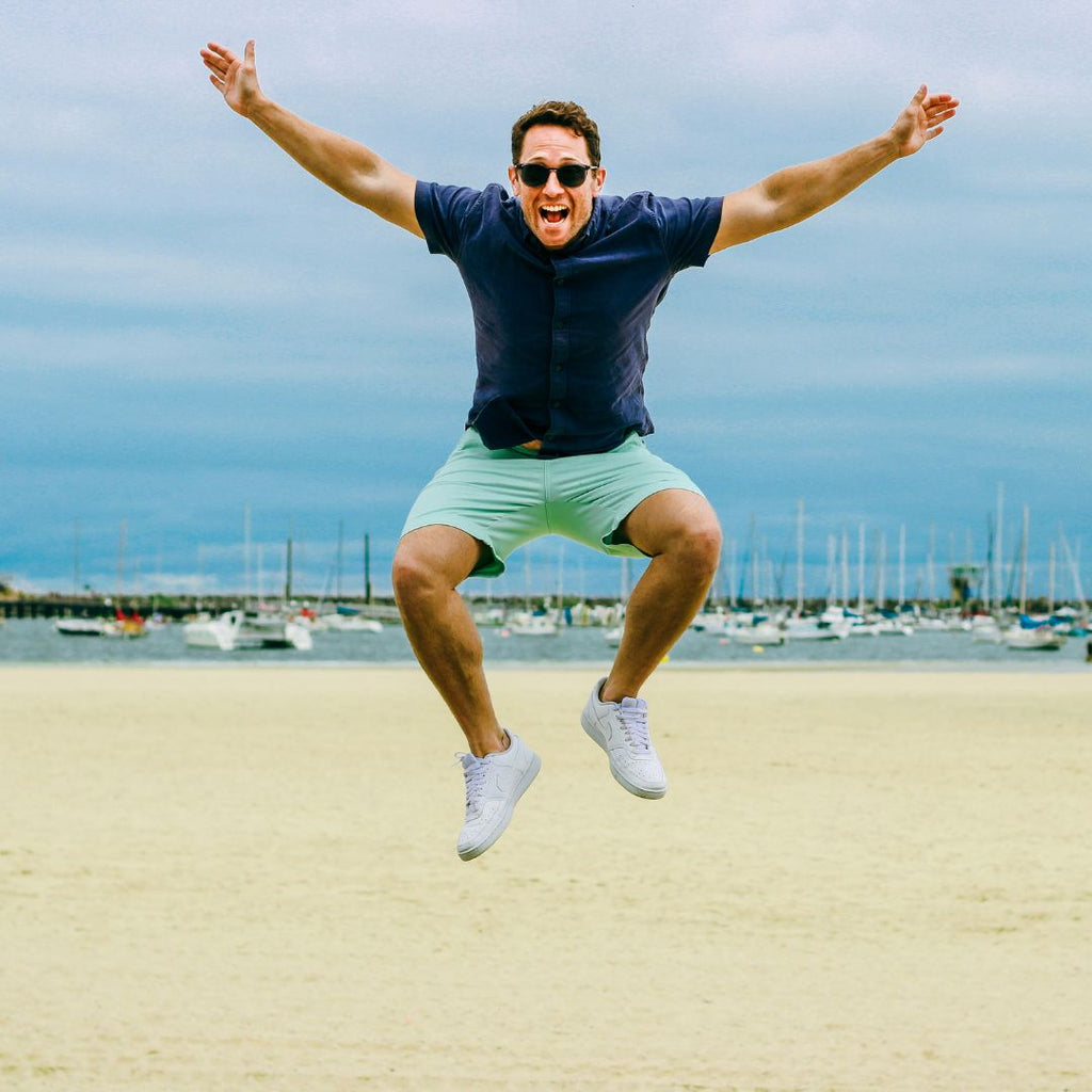 A man wearing a dark shirt, Frankster's light green men's shorts, and sunglasses is jumping in the air on a sandy beach with a joyful expression. The beach has several boats anchored in the background under a cloudy sky, perfectly capturing the essence of summer breeze attire.