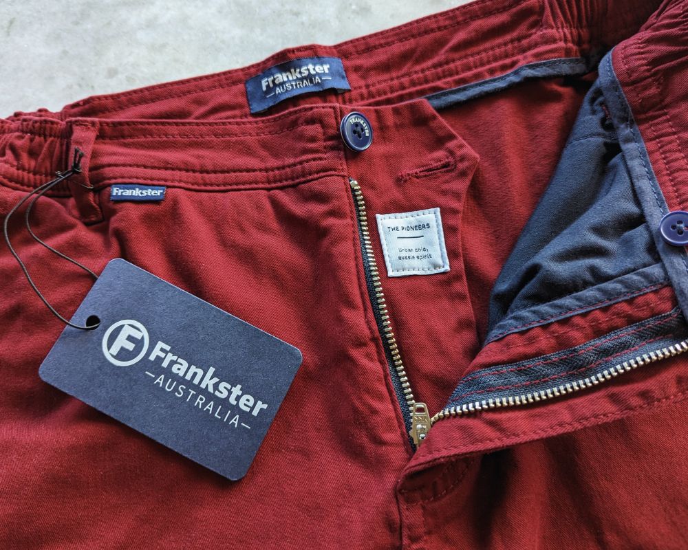 Close-up of red shorts with an open zipper and visible blue fabric lining. The pants have a tag attached with the brand name "Frankster Australia" and another small white tag with product information sewn inside.