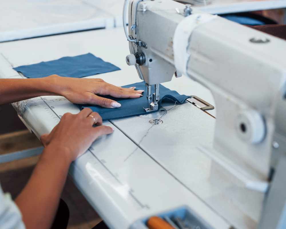 Close-up of a person using a sewing machine on a light-colored table. They are sewing a piece of denim fabric, guiding it with both hands. Various sewing tools are scattered around, and the background shows other tables and workstations.