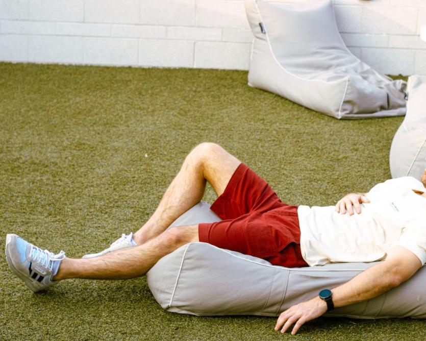 A man in a white shirt, red frankster men shorts, and a white cap lies on a wooden bench in the park. He has his right arm resting on his chest and wears reflective sunglasses. The sun is shining brightly, casting shadows, and the grass around is green and lush—a typical Frankster enjoying the day with "The Pioneers.