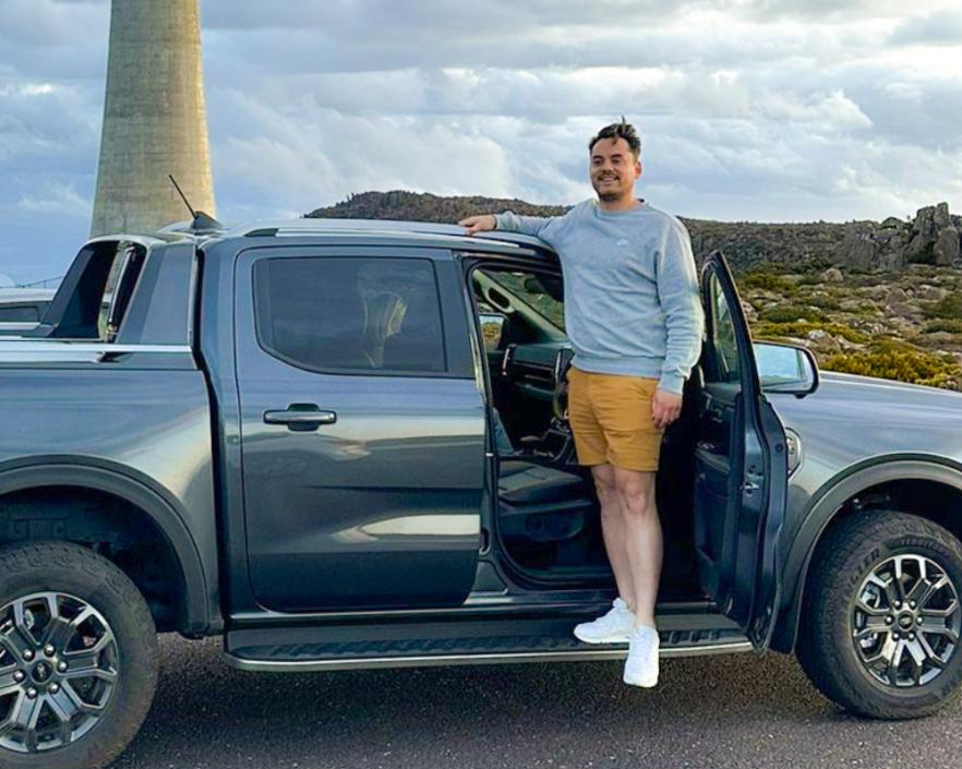 A man in a gray sweatshirt and dark yellow frankster shorts stands on the footboard of a gray pickup truck with the driver's side door open. He is smiling and leaning on the roof of the truck. In the background, there is a large tower and a cloudy sky.