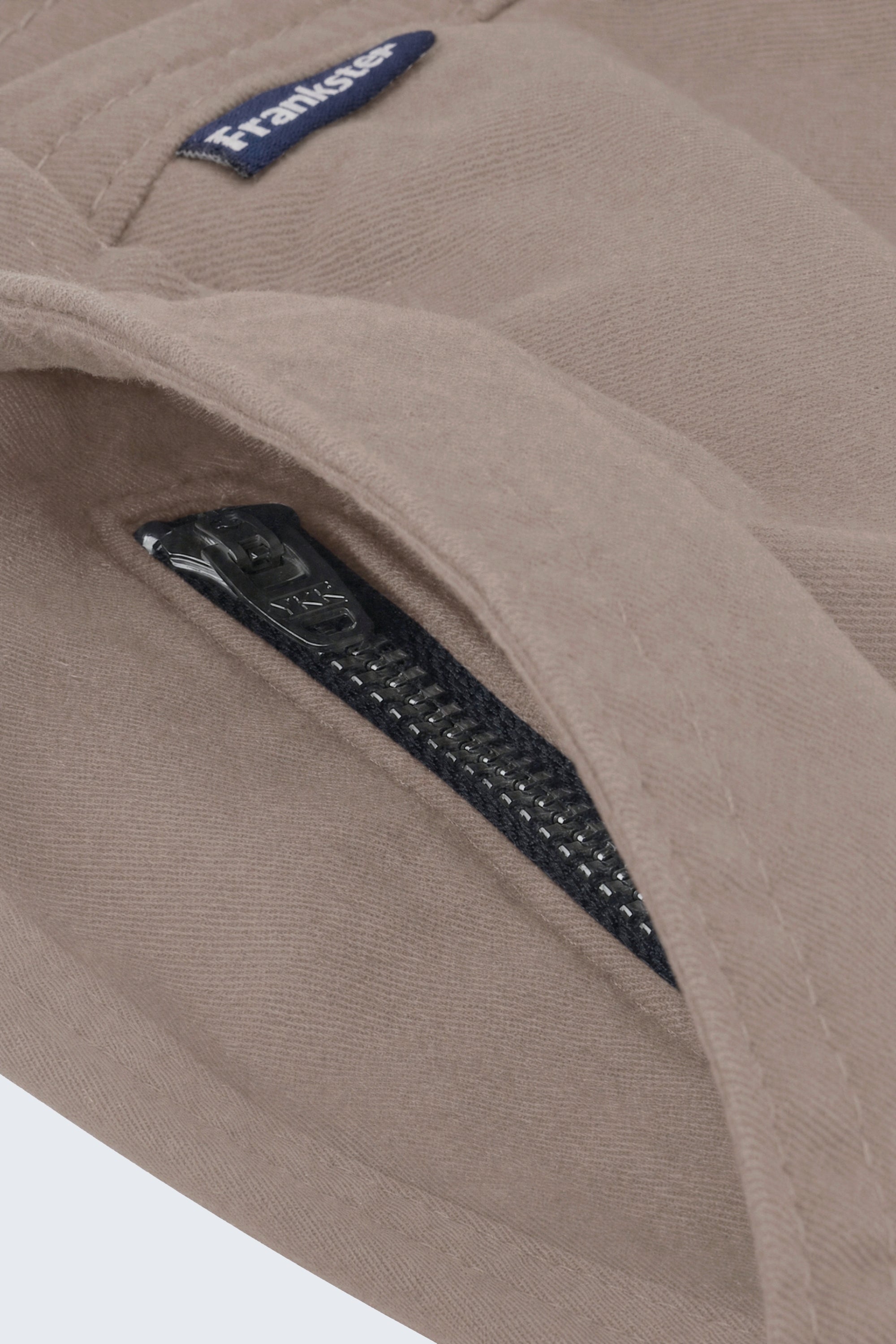 Close-up of beige men's shorts fabric showing a black zipper pocket and a small navy Frankster label, highlighting the durable material of Nomads * The Everydays men's shorts by Frankster.