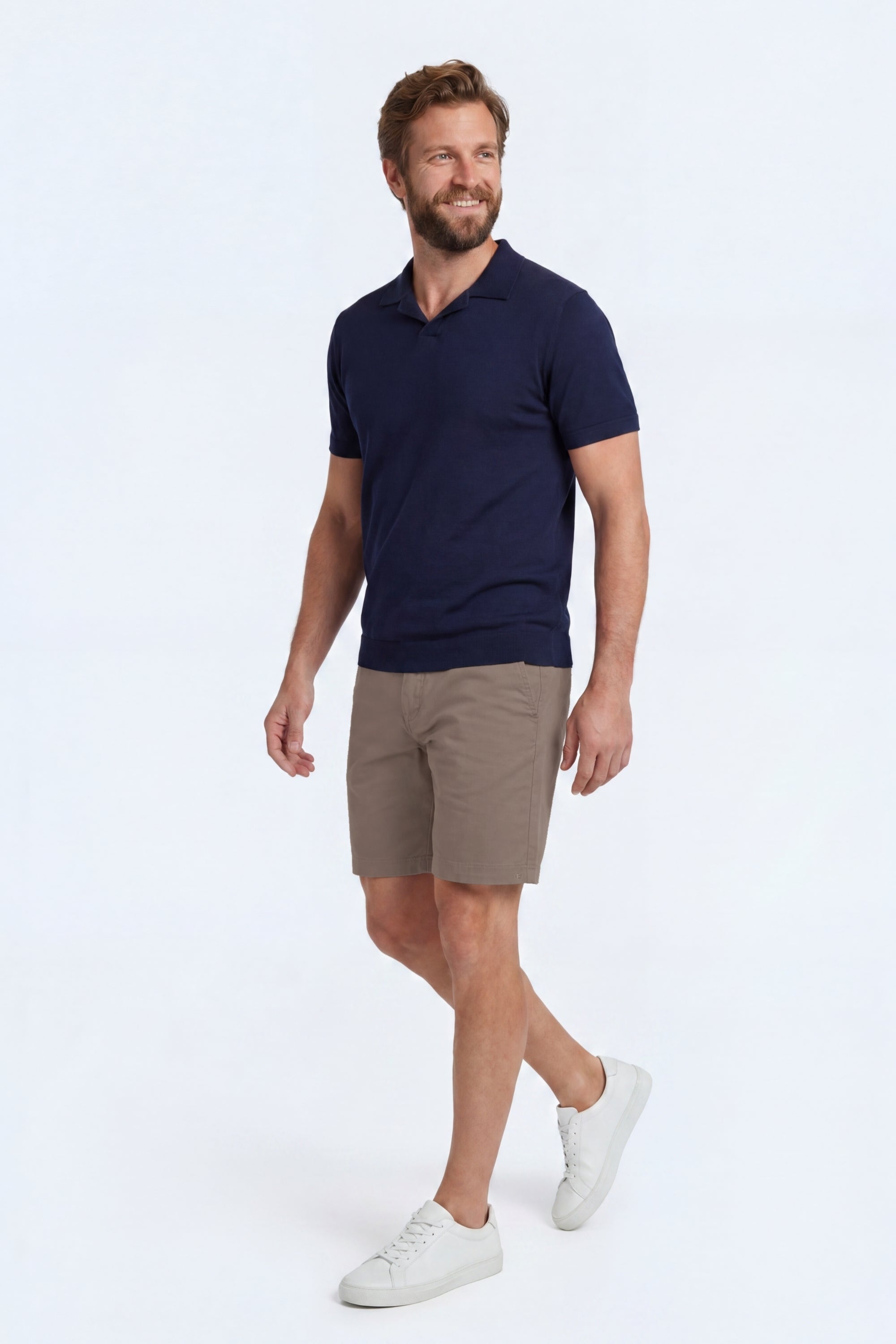 A smiling man wearing beige cotton blend men's shorts from Frankster’s Nomads * The Everydays collection paired with a navy blue polo and white sneakers, showcasing comfortable, stylish shorts for everyday wear.