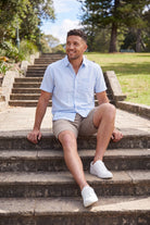 A smiling man wearing light blue casual shirt and beige Nomads * The Everydays men's shorts by Frankster, made from breathable fabric, sitting outdoors on stone steps.