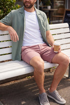 Man sitting on a bench wearing dusty pink Sundowners * The Everydays men's shorts by Frankster, paired with a white t-shirt and green shirt, holding a coffee cup.