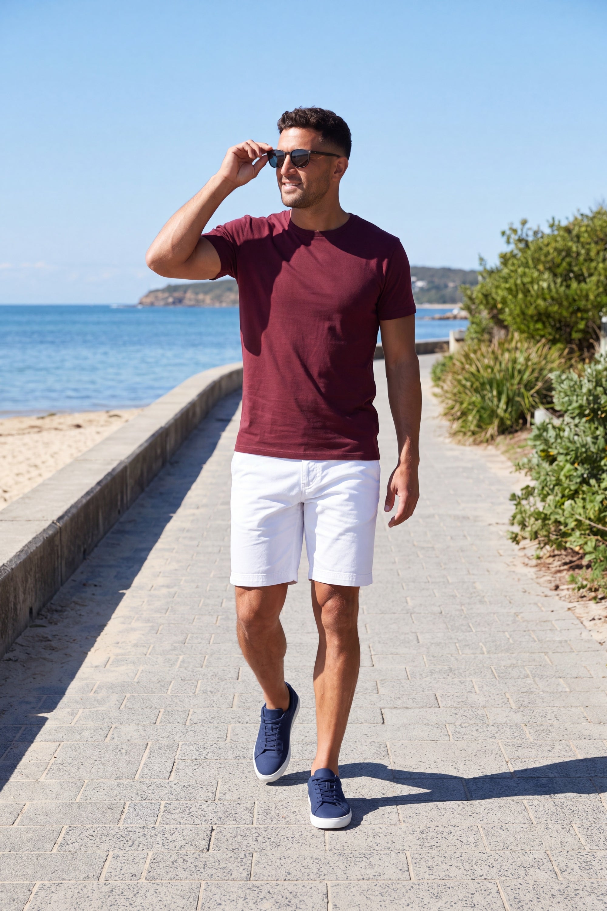 Man wearing Whitecaps * The Everydays white men's shorts by Frankster, paired with a burgundy shirt and navy sneakers on a sunny beach path.