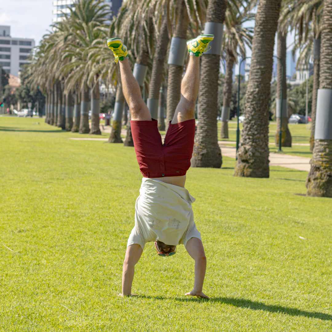 A person in a white shirt and red frankster shorts performs a handstand on a grassy lawn lined with tall palm trees on a sunny day. Buildings are visible in the background.